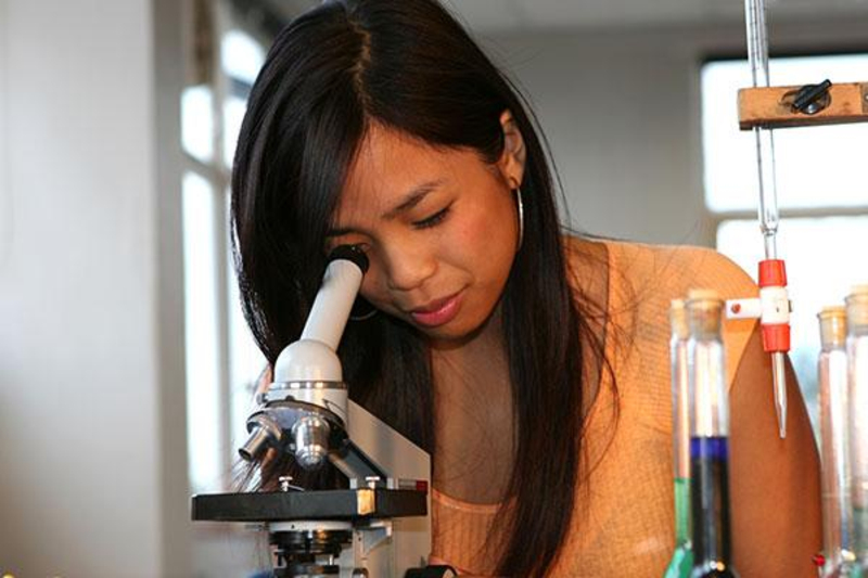 Eine junge Frau mit langen schwarzen Haaren schaut konzentriert in ein Mikroskop. Neben ihr stehen Reagenzgläser mit bunten Flüssigkeiten. 