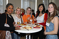 Fünf Frauen stehen an einem Stehtisch und tauschen sich aus. Vor ihnen stehen Getränke und befüllte Teller mit Essen. 