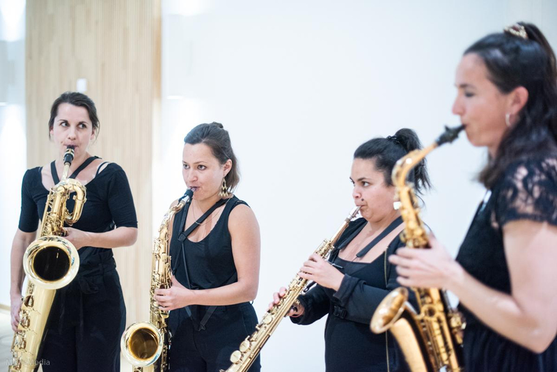 Vier schwarz gekleidete Frauen stehen im Halbkreis und spielen Saxophon.