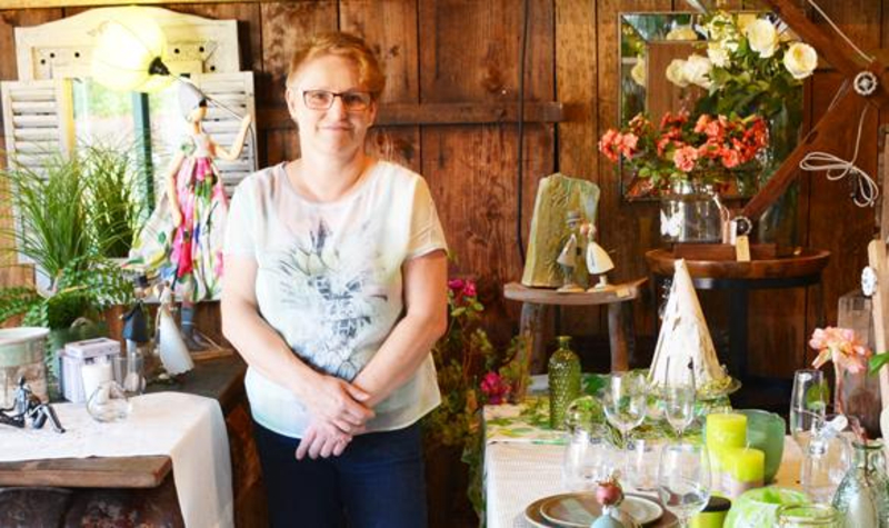 Eine Frau mit kurzen blonden Haaren und Brille steht in einem gemütlich wirkenden Verkaufsraum. Dieser ist mit Vasen, Blumen und Keramikartikeln gefüllt.