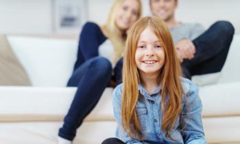 Mädchen in Jeanshemd und mit roten Haaren lächelt in die Kamera. Im Hintergrund sitzen die Eltern auf einer Couch.