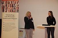 Die zwei Frauen stehen nebeneinander auf dem Podium. Frau Schmidt von der Kontaktstelle Ravensburg hält ein Mikrofon in der Hand. Sie wird von Frau Pantel interessiert angeschaut.