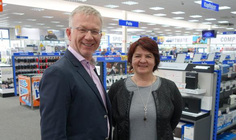 Ein Mann und eine Frau stehen nebeneinander in einem Elektrofachmarkt und schauen freundlich in die Kamera. Der Mann trägt einen Anzug mit rosa Hemd, die Frau hat rote Haare und eine schwarze Jacke an.