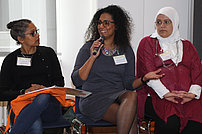 Drei Teilnehmerinnen des Mentorinnen-Programms sitzen nebeneinander auf Stühlen. Die Dame in der Mitte hält ein Mikrofon in der Hand und erzählt. Sie wird interessiert angeschaut.