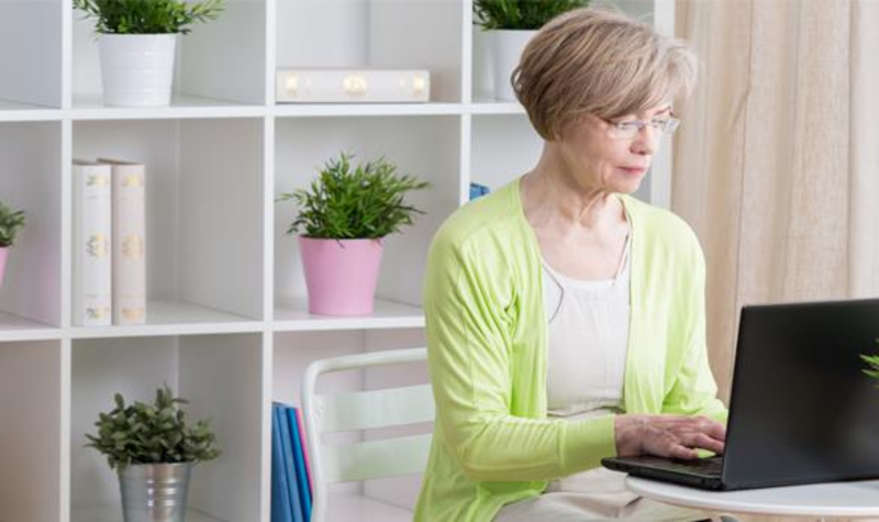 Eine Frau mittleren Alters sitzt vor einem Regal mit vielen Pflanzen am Laptop und arbeitet.