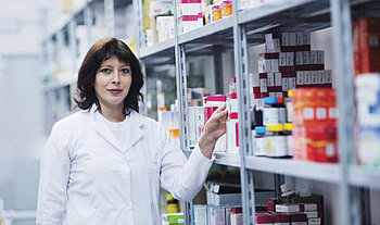 Eine Frau in weißem Kittel steht an ein Regal gelehnt. Das Regal ist mit Medikamenten gefüllt.