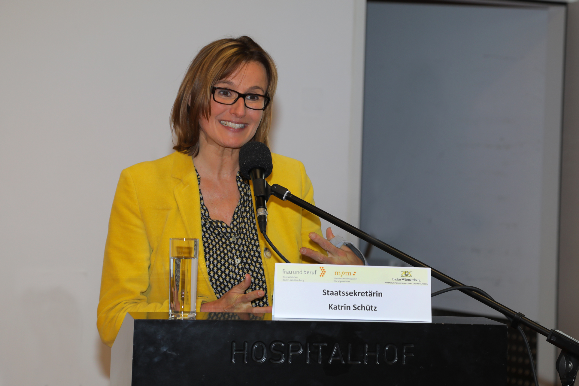 Staatssekretärin Katrin Schütz steht auf dem Podium und spricht in ein Mikrofon. Sie trägt einen gelben Blazer.