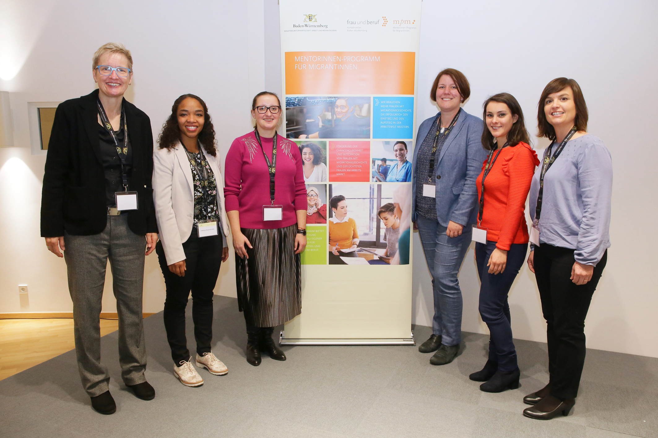 Sechs Beteiligte am Mentorinnen-Programm für Migrantinnen stehen in einem Halbkreis vor einem Roll-up des Landesprogramms Kontaktstellen Frau und Beruf.
