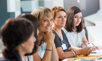 Fünf Frauen sitzen in einer Reihe an einem Konferenztisch. Alle schauen interessiert geradeaus.