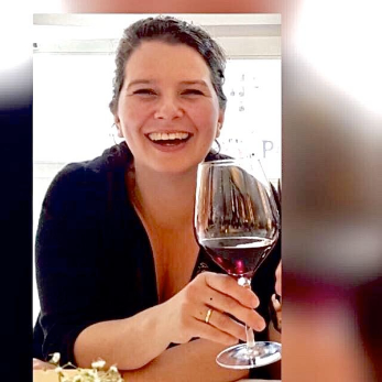 Brünette Frau mit zurückgebundenem Haar hält ein Glas mit Rotwein in der Hand und lacht herzlich und offen in die Kamera
